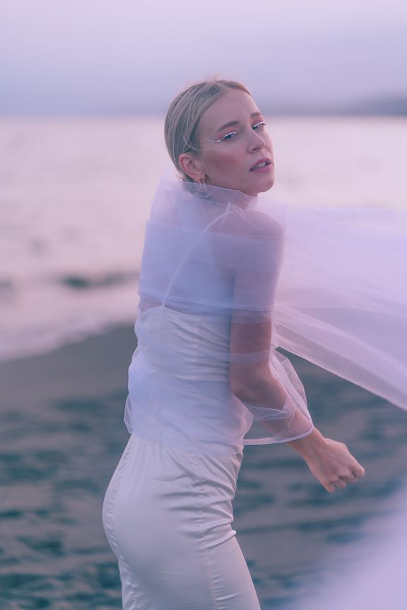 Blonde woman in white outfit wrapped in sheer tulle veil poses on beach at dusk.