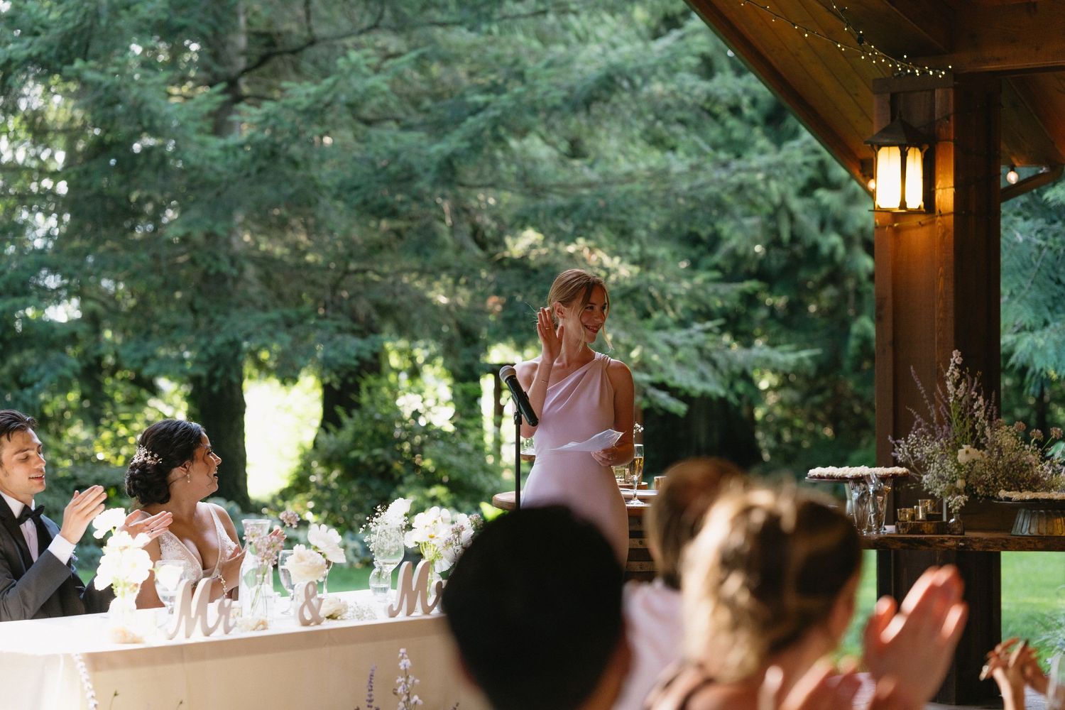 Bridesmaid in lavender dress giving a speech at outdoor wedding reception under a wooden pavilion.