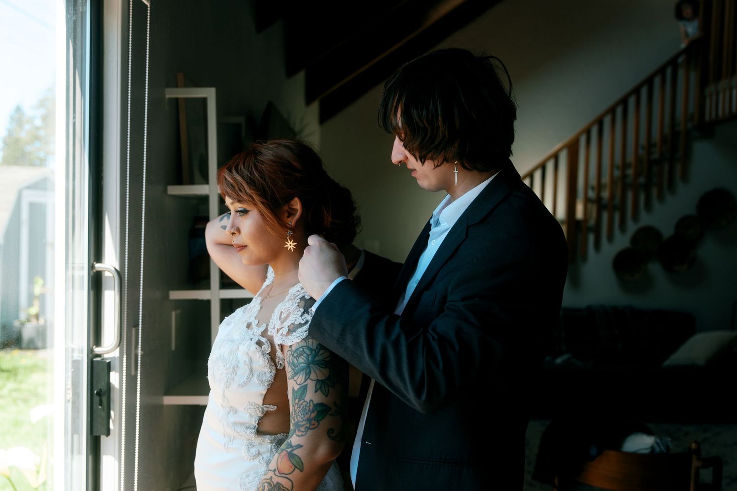 Groom helping bride with jewelry near window with staircase in background.