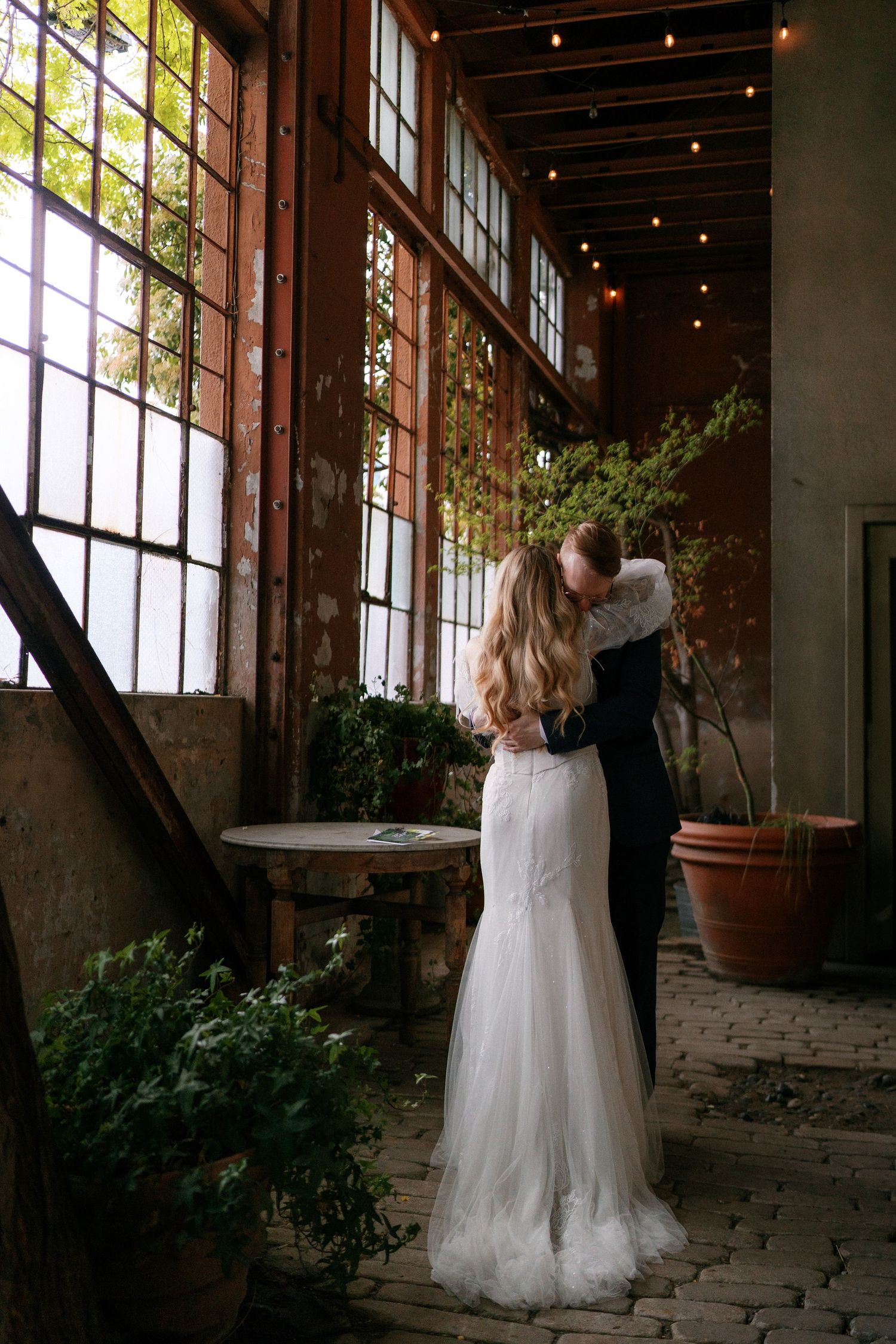 Bride and groom share first dance embrace in a romantic industrial venue with string lights.