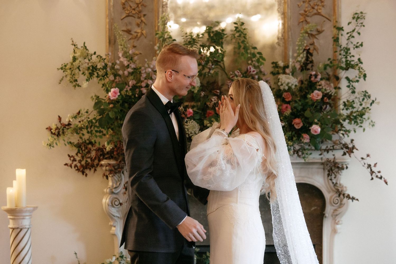 Bride tenderly touches groom's face before romantic floral fireplace at candlelit wedding ceremony.