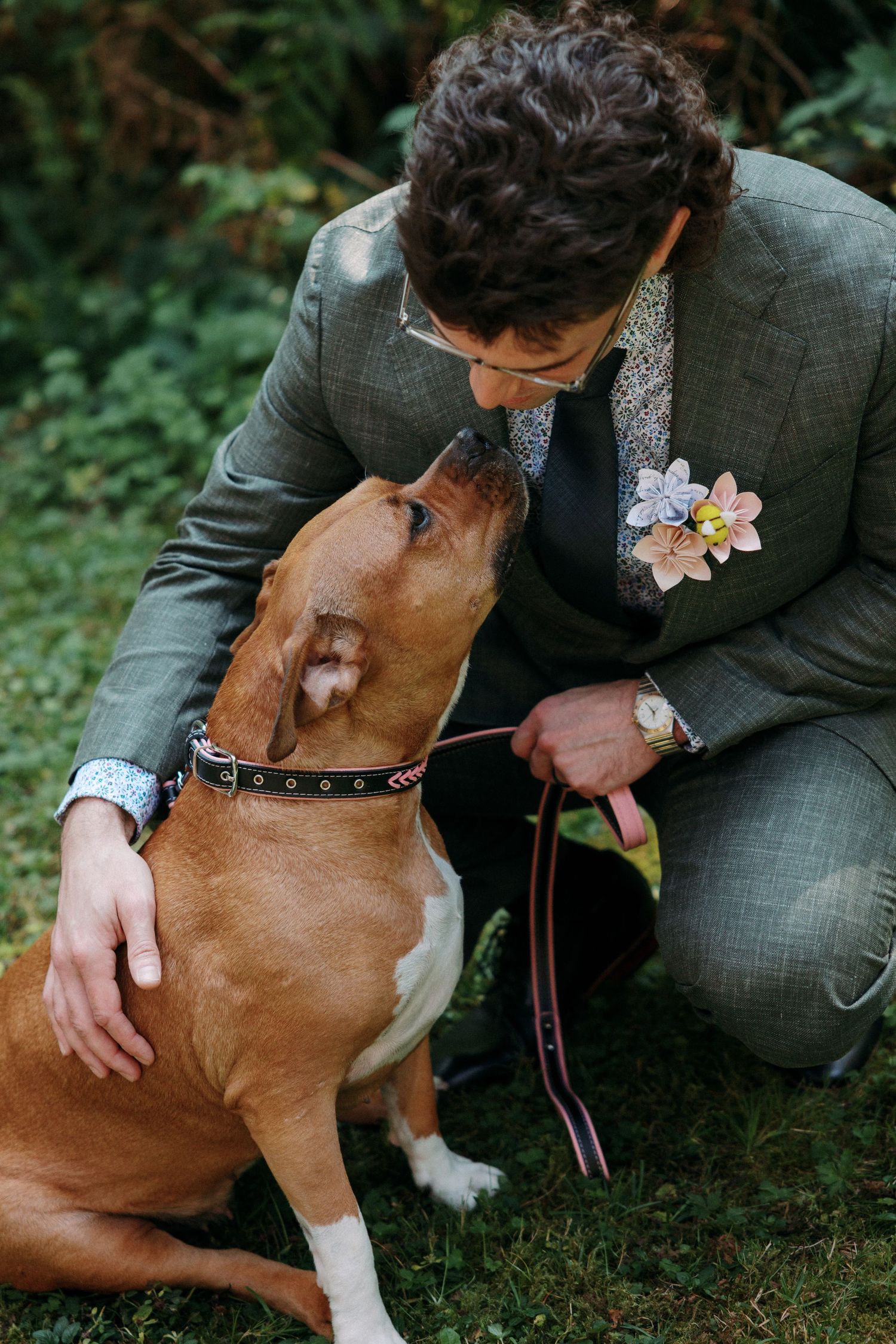 A groom in a gray suit crouches to interact with his brown and white dog on a green lawn.