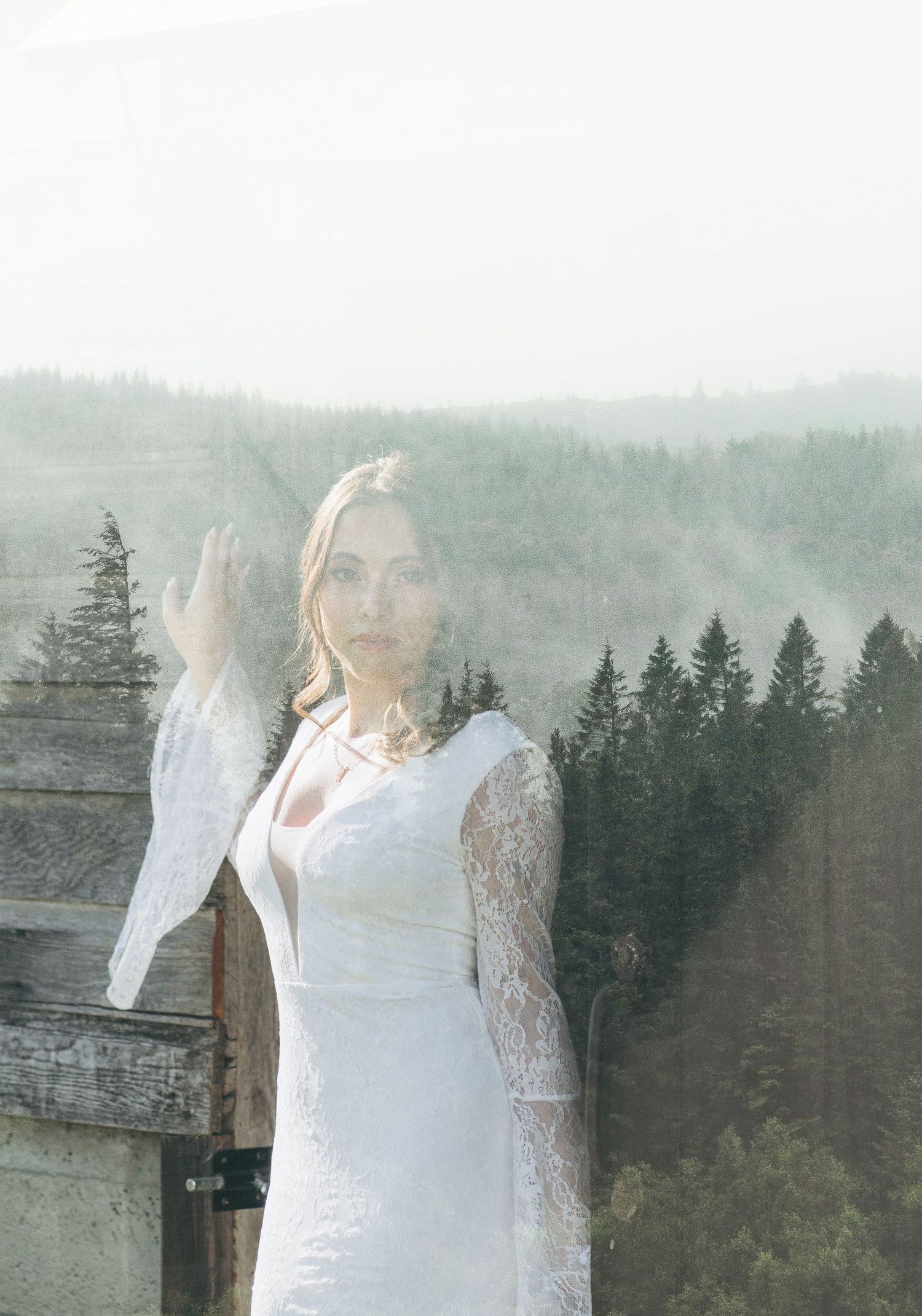 Double exposure portrait of bride in white lace dress blended with misty forest landscape.