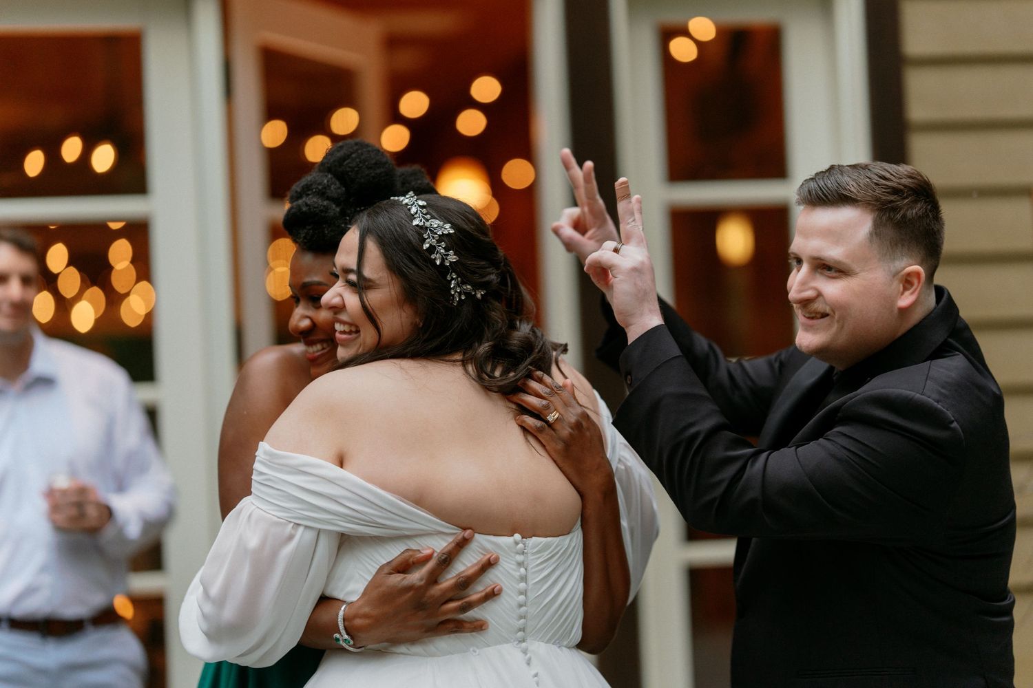Bride in white gown hugging guest while groom flashes peace signs at wedding reception.