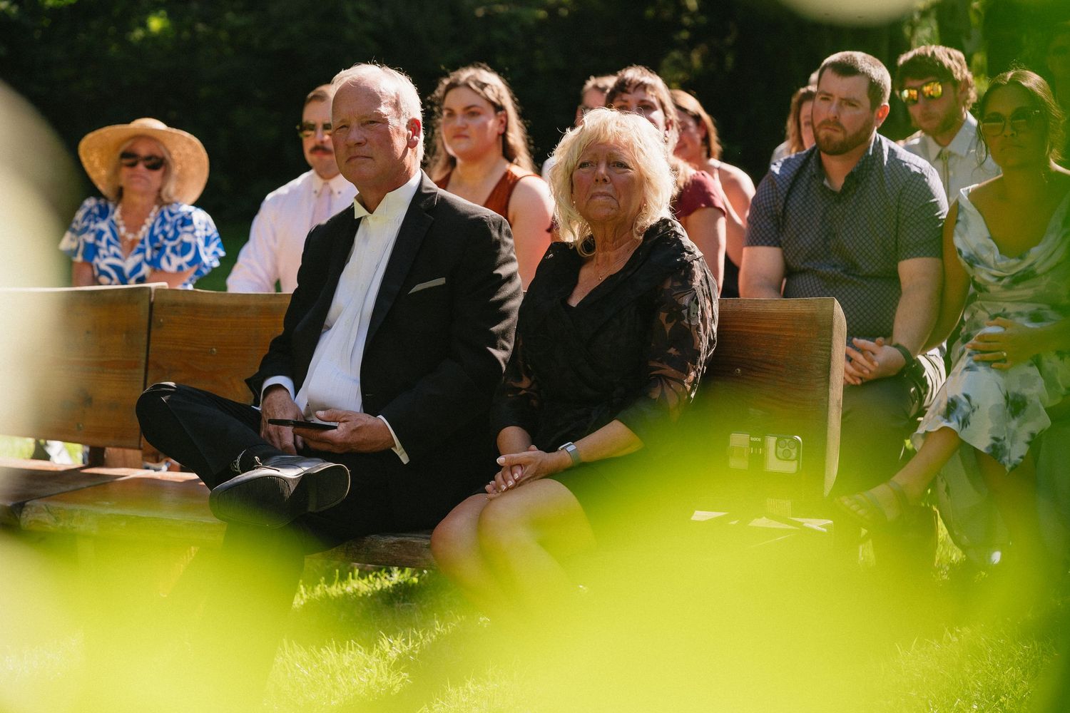 Wedding guests seated on benches watching outdoor ceremony, couple in foreground wearing black attire.