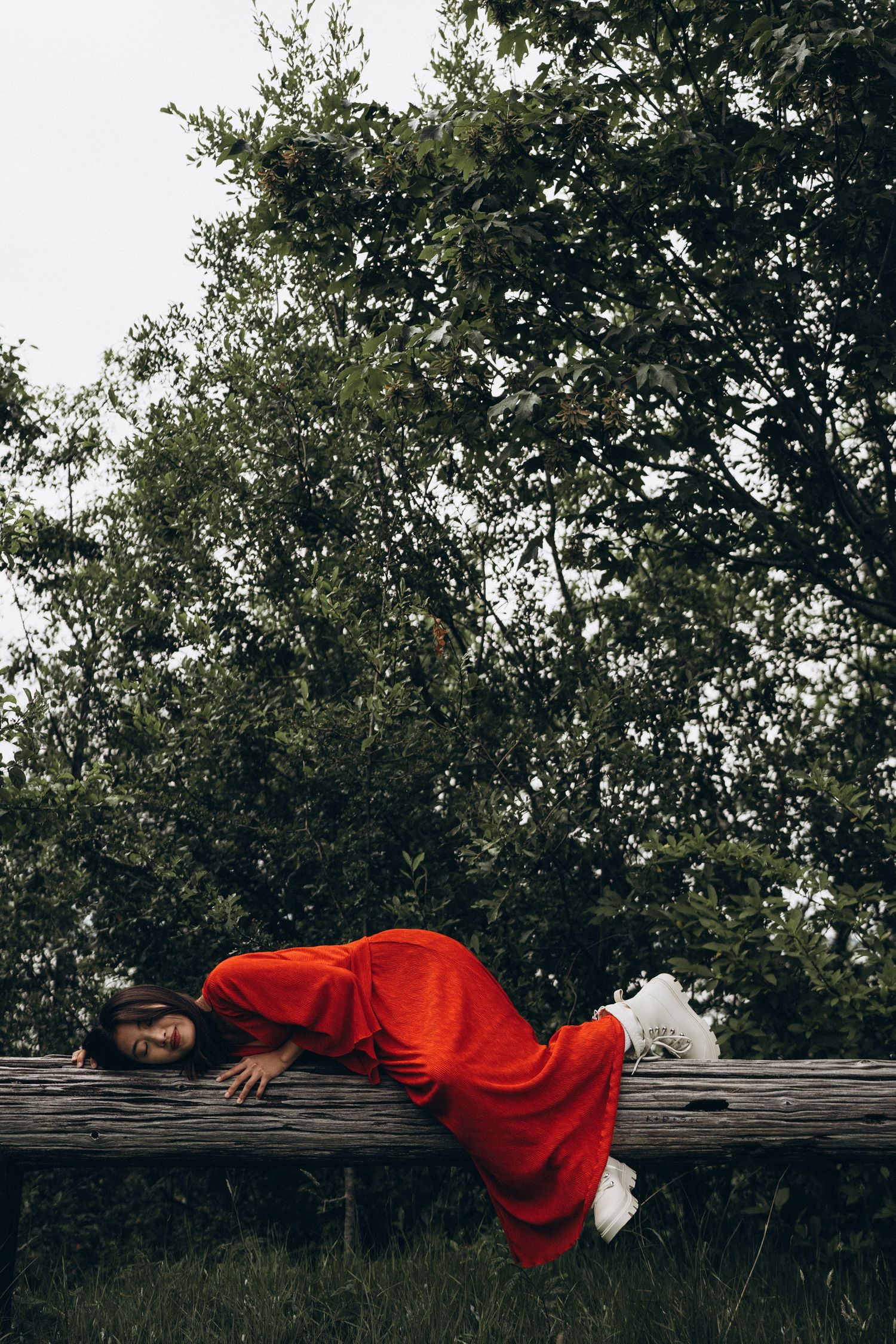 Woman in red dress lying on a rustic wooden fence surrounded by lush green trees.