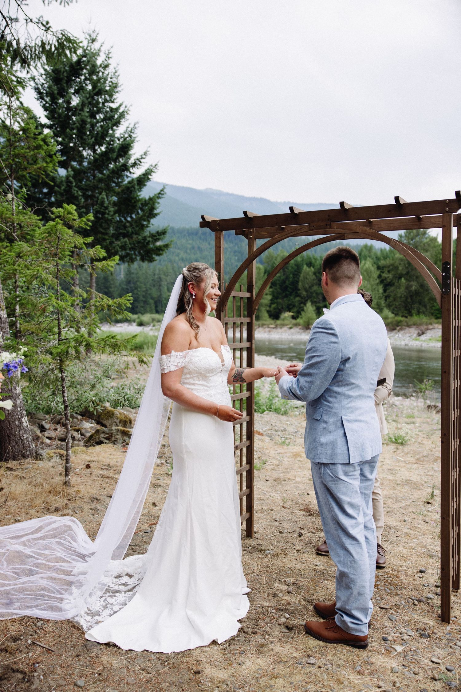 Bride in off-shoulder gown with flowing veil holds hands with groom in blue suit at outdoor ceremony.
