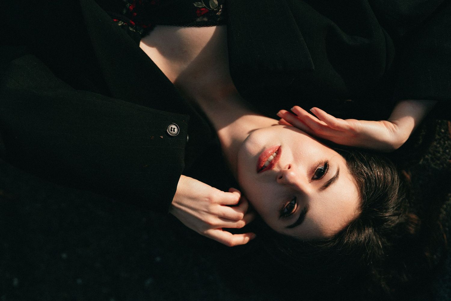 Close-up portrait of young woman lying down in sunlight, hand near face, wearing black jacket.