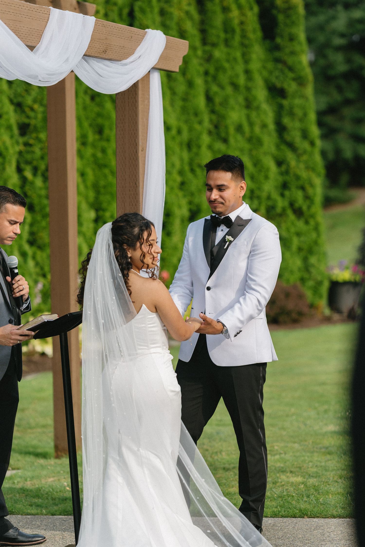 Radiant bride and groom hold hands and smile while exchanging vows under a wooden pergola outdoors.