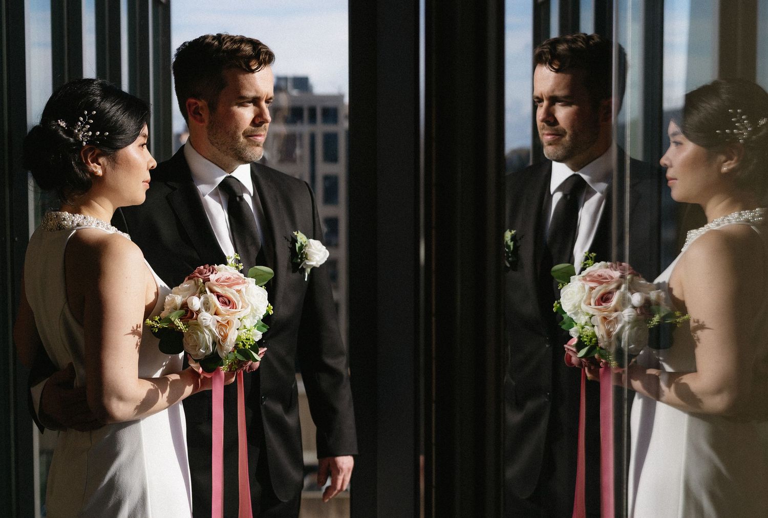 Bride and groom standing by tall windows with romantic reflection, bride holding pink and white wedding bouquet.