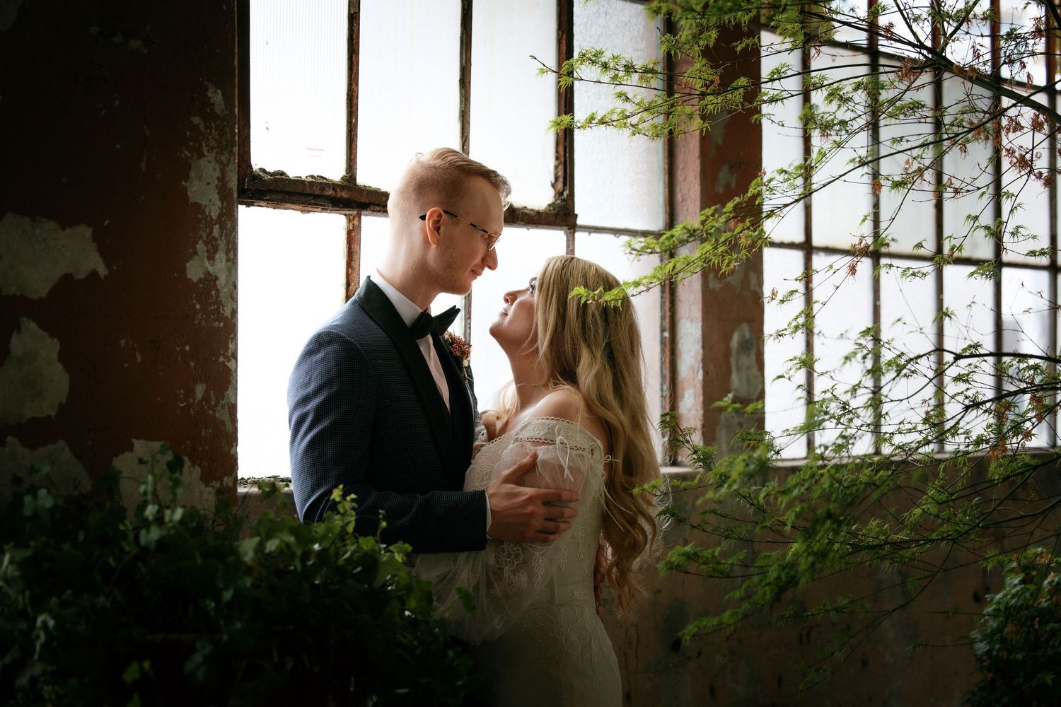 Bride and groom sharing a romantic moment by a window surrounded by lush greenery.
