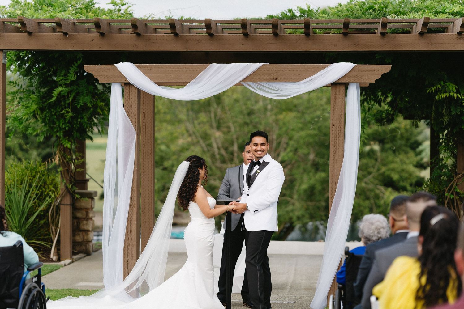 Smiling bride and groom exchange vows under a white-draped wooden pergola at an elegant outdoor wedding ceremony.