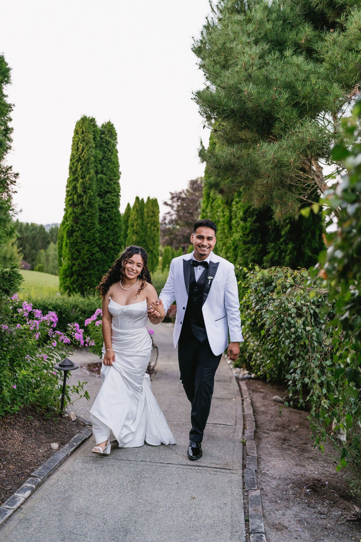 Happy bride and groom walk along a garden path lined with cypress trees and pink flowering bushes.