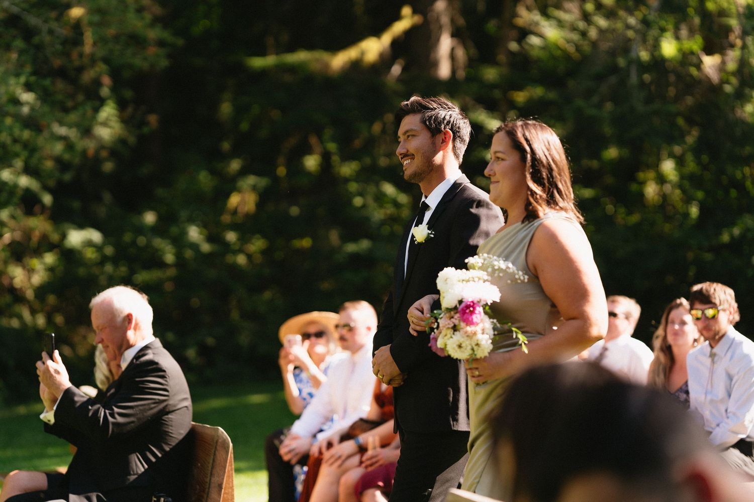 Groomsman and bridesmaid walk down sunlit outdoor wedding ceremony aisle holding floral bouquet.