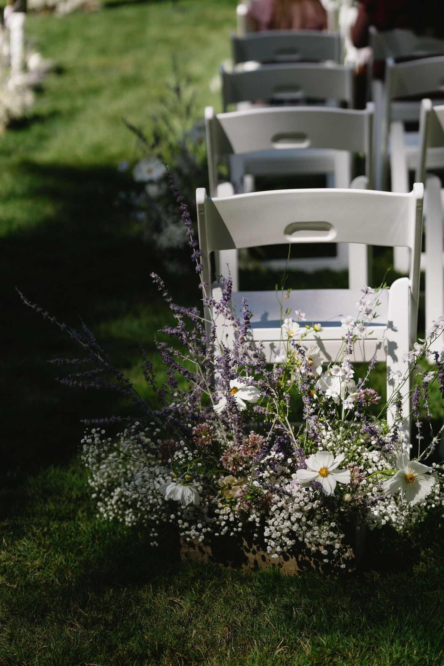 Wildflower arrangement with lavender and daisies decorating white chairs at outdoor wedding ceremony.