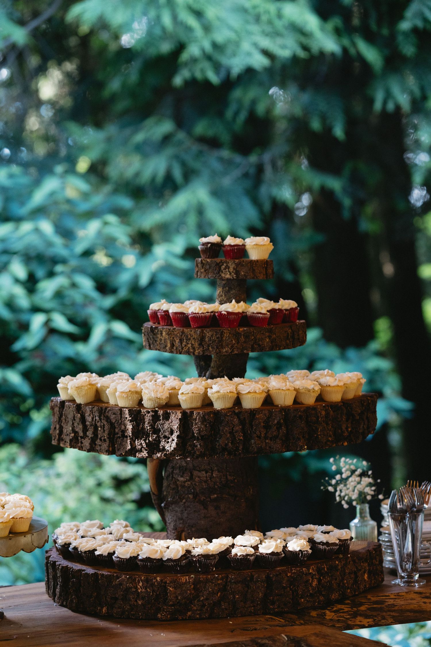 Elegant four-tier rustic wood slab cupcake tower display with white and red frosted cupcakes outdoors.