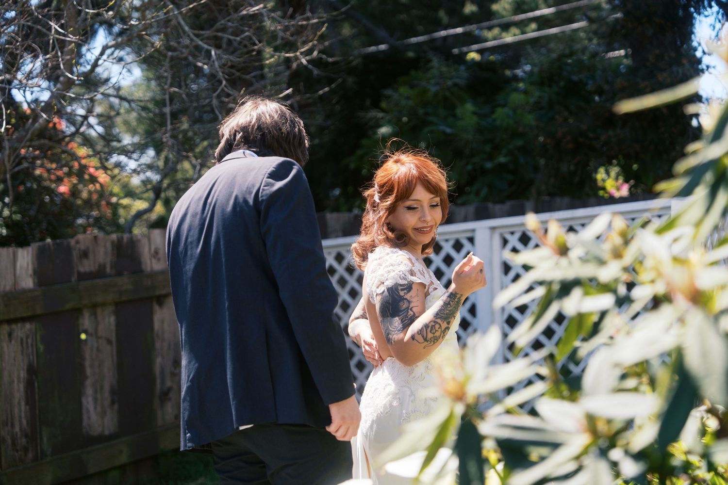 Bride in lace wedding dress looks back over shoulder at groom in garden setting.