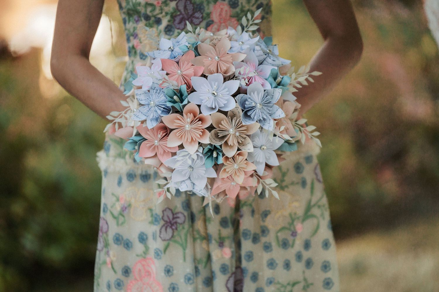 Bridesmaid in floral dress holds handcrafted paper flower bouquet in soft pastel pink and blue tones.
