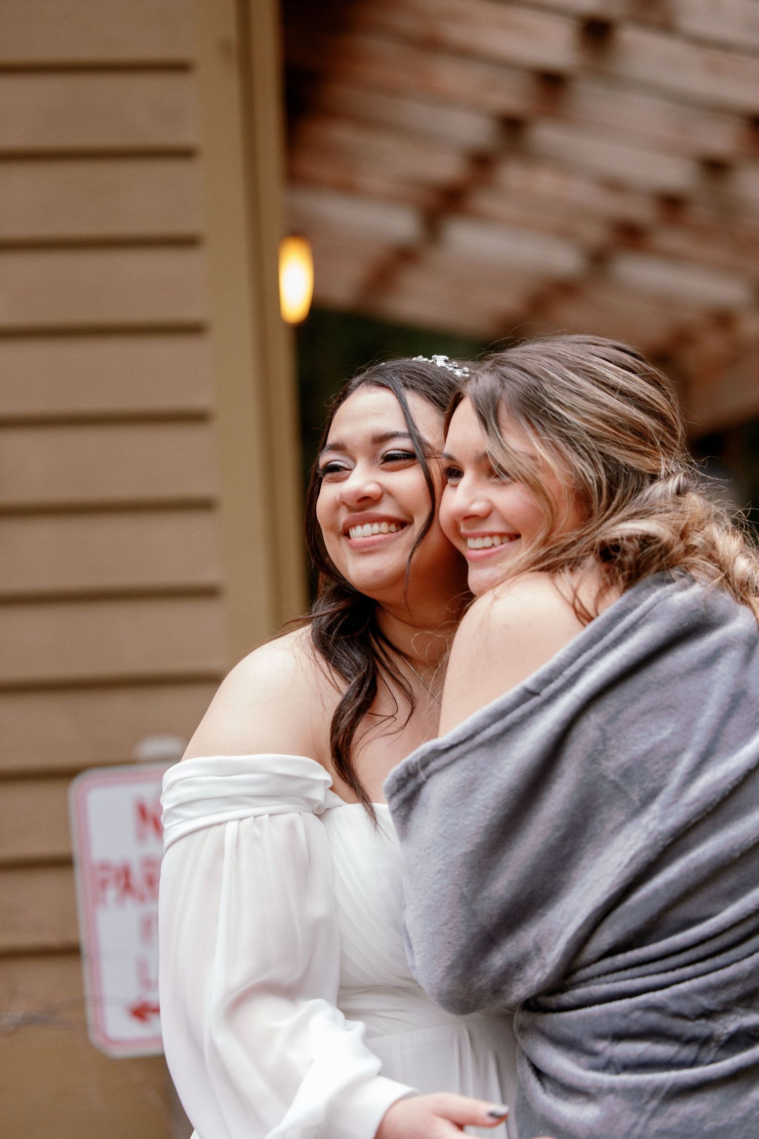 Bride in white off-shoulder gown embracing a guest in a gray wrap at outdoor wedding.