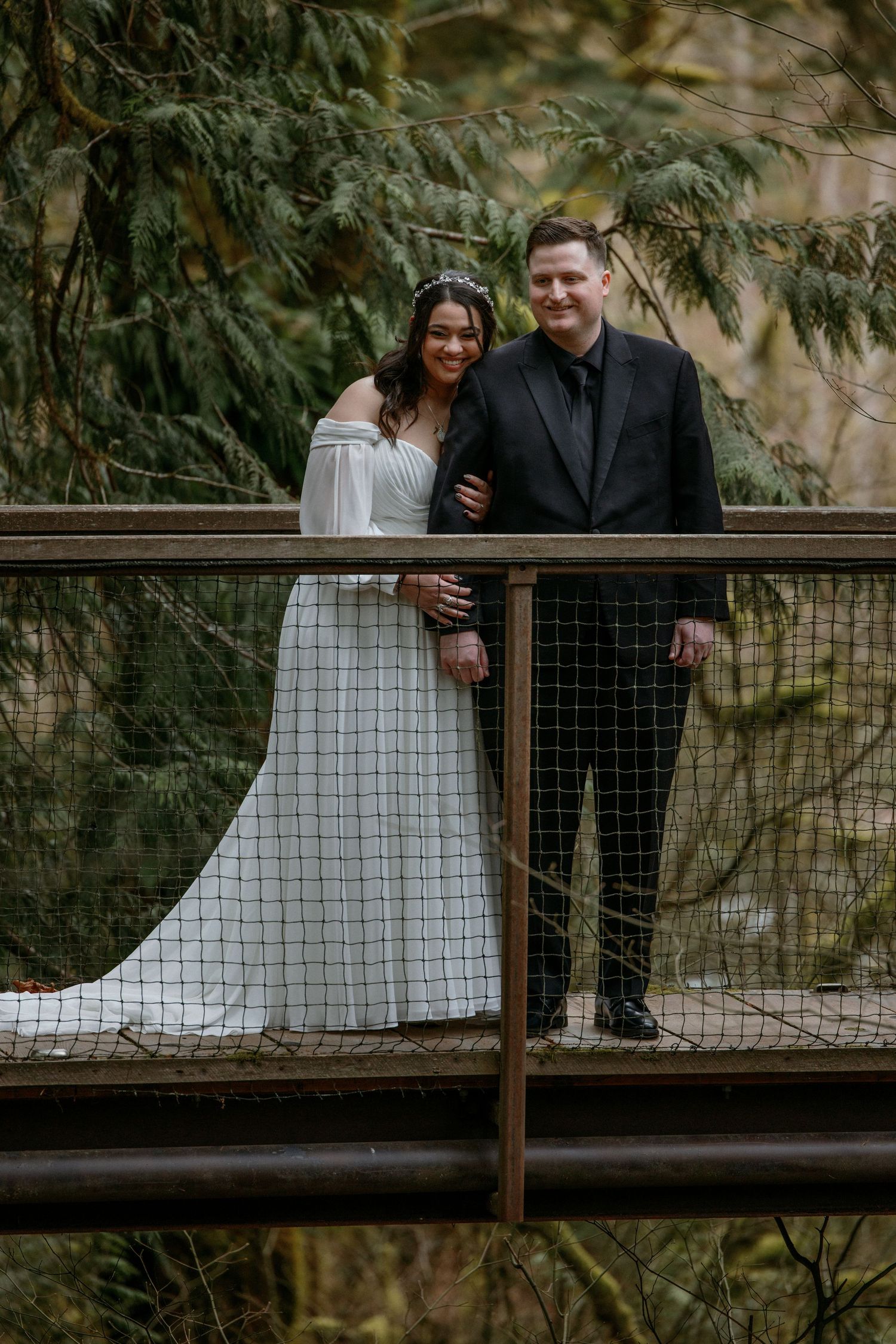 Smiling bride and groom pose together on wooden bridge surrounded by lush evergreen forest trees.