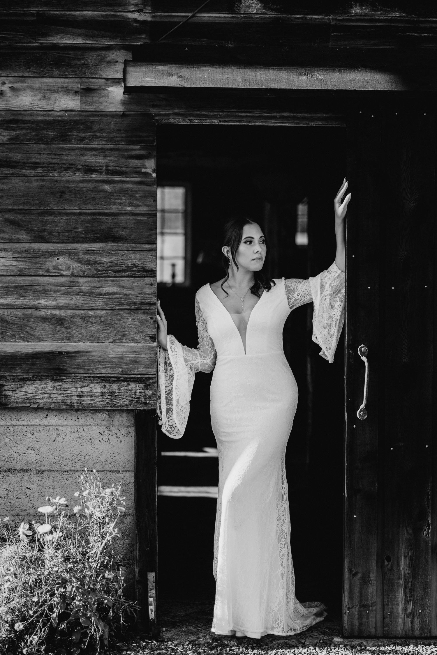Black and white portrait of bride in fitted lace wedding gown posing in rustic barn doorway.