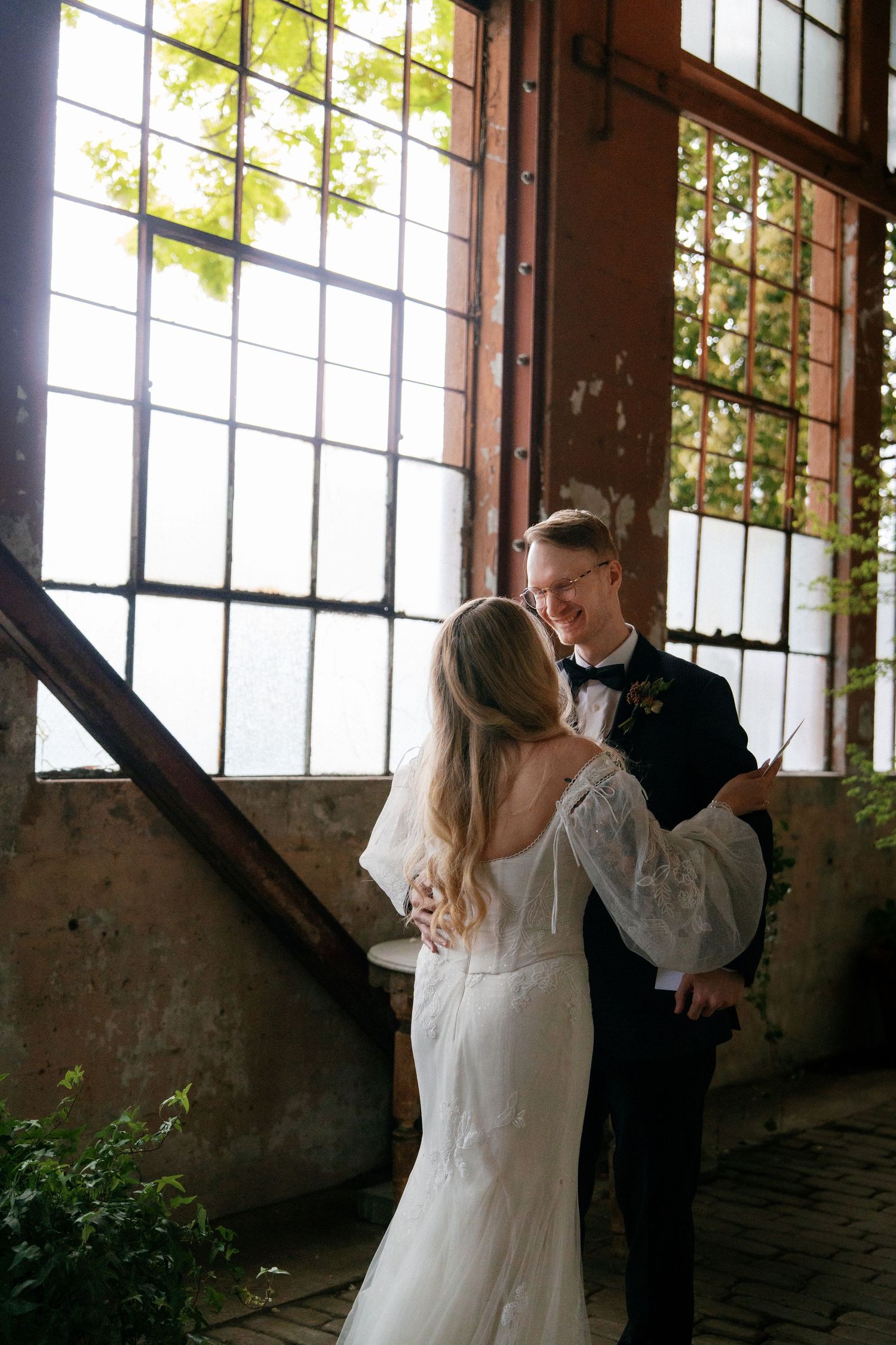 Bride and groom share an intimate moment near industrial windows at their wedding venue.