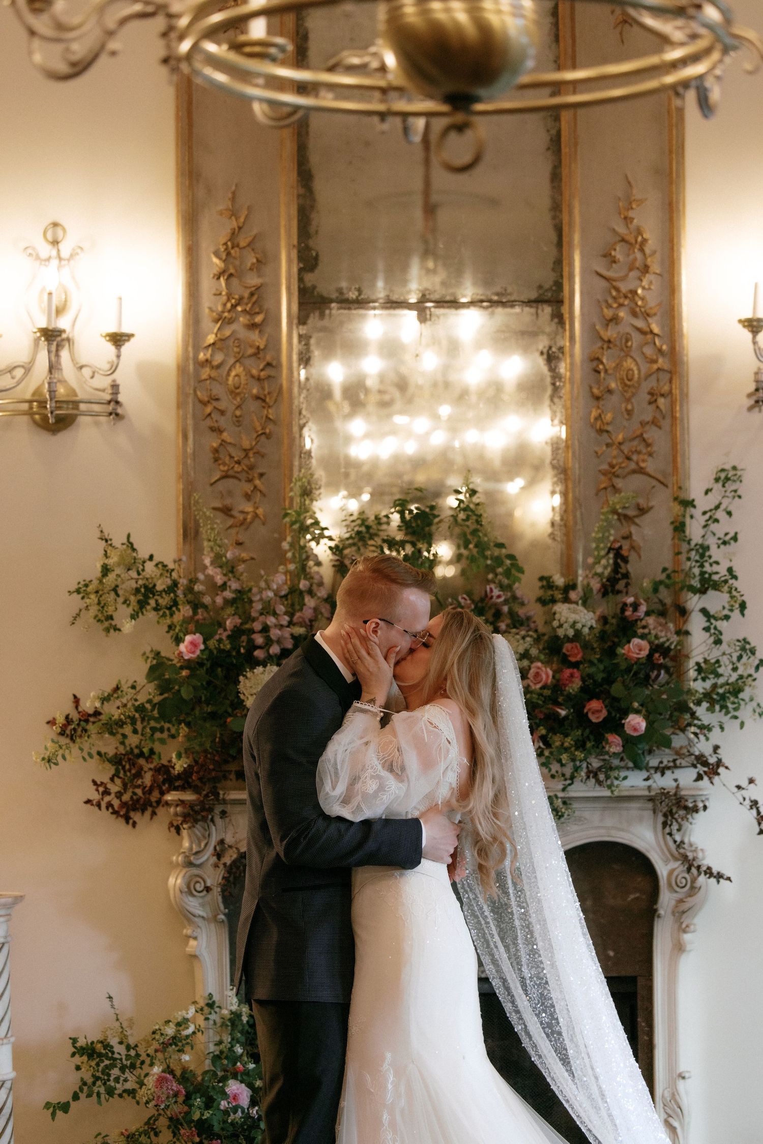 Newlyweds share romantic first kiss beneath chandelier before floral fireplace at elegant wedding venue.