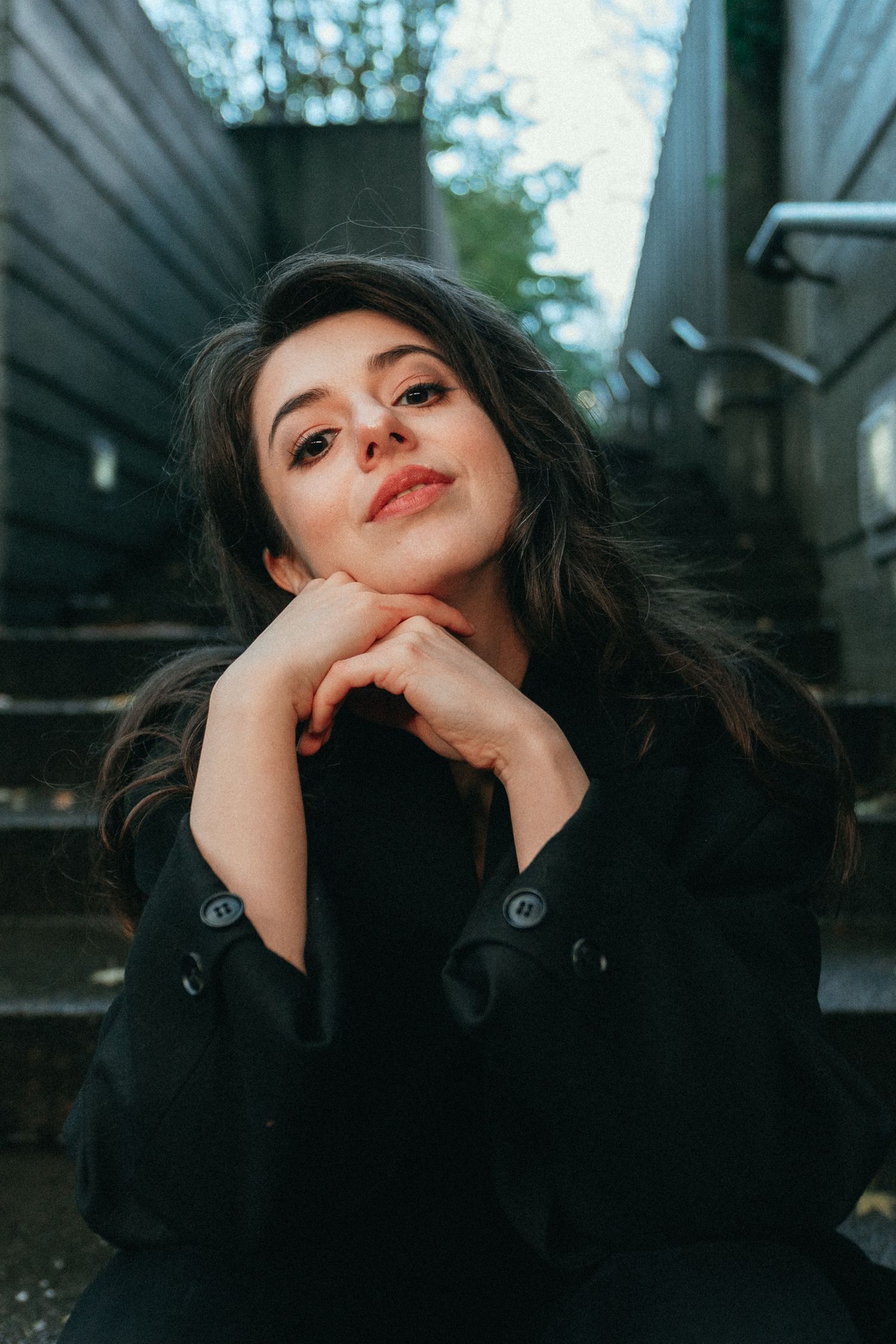 Smiling young brunette woman in black coat sits on steps with hands clasped under chin.