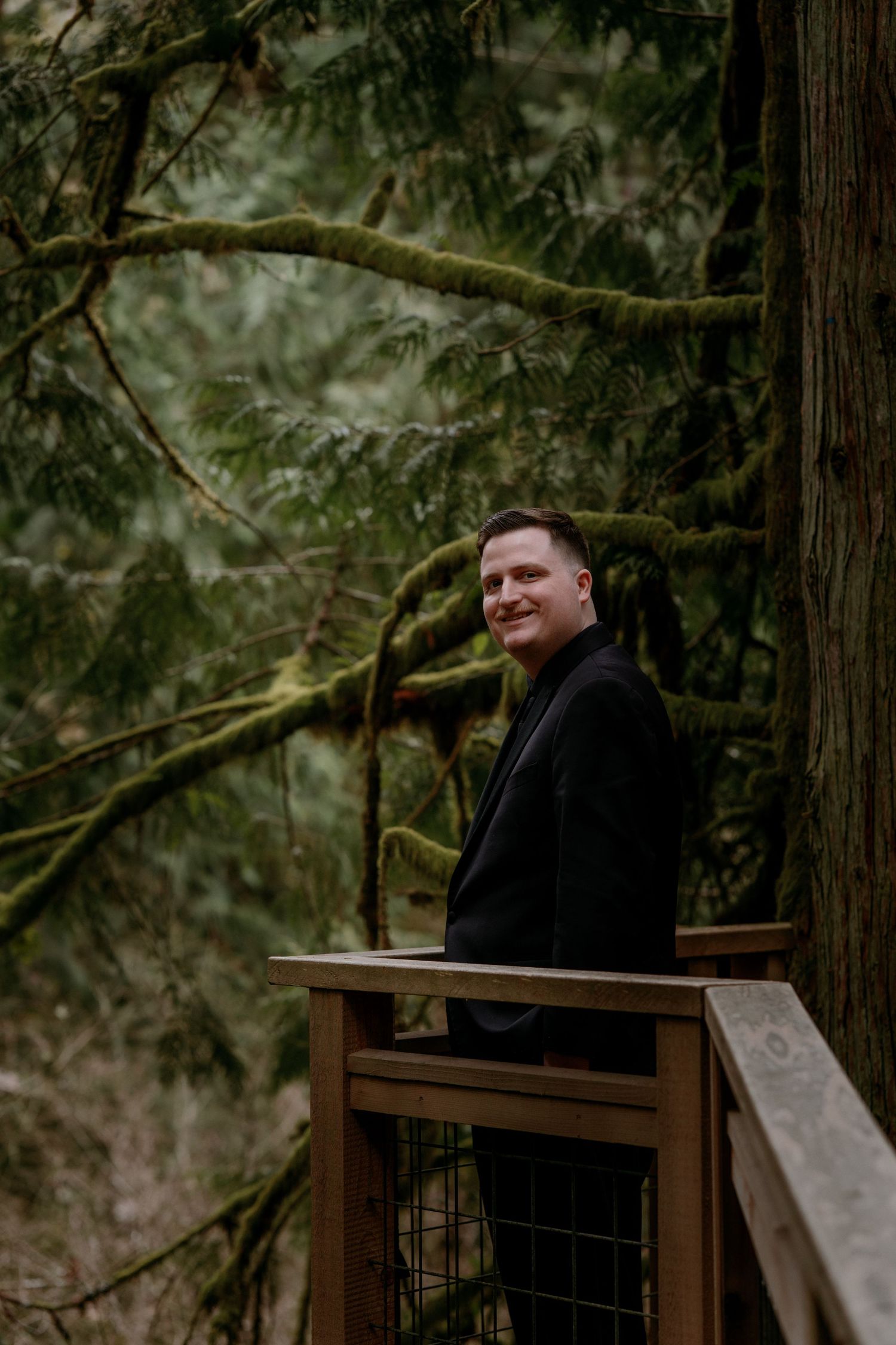 Smiling groom in black suit leans on wooden railing surrounded by moss-covered trees in forest.
