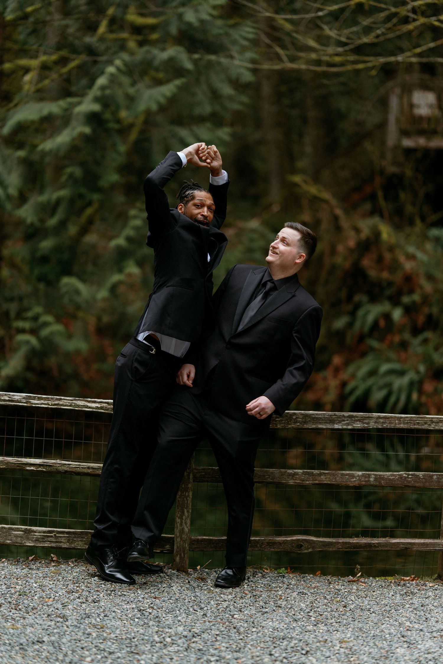 Two grooms in black suits posing playfully by a wooden fence in a forest wedding.