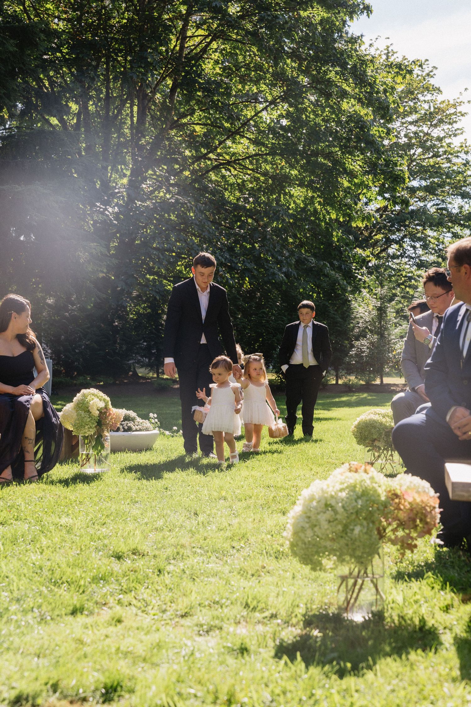 Flower girls and groomsman walk down outdoor wedding aisle lined with hydrangea floral arrangements.