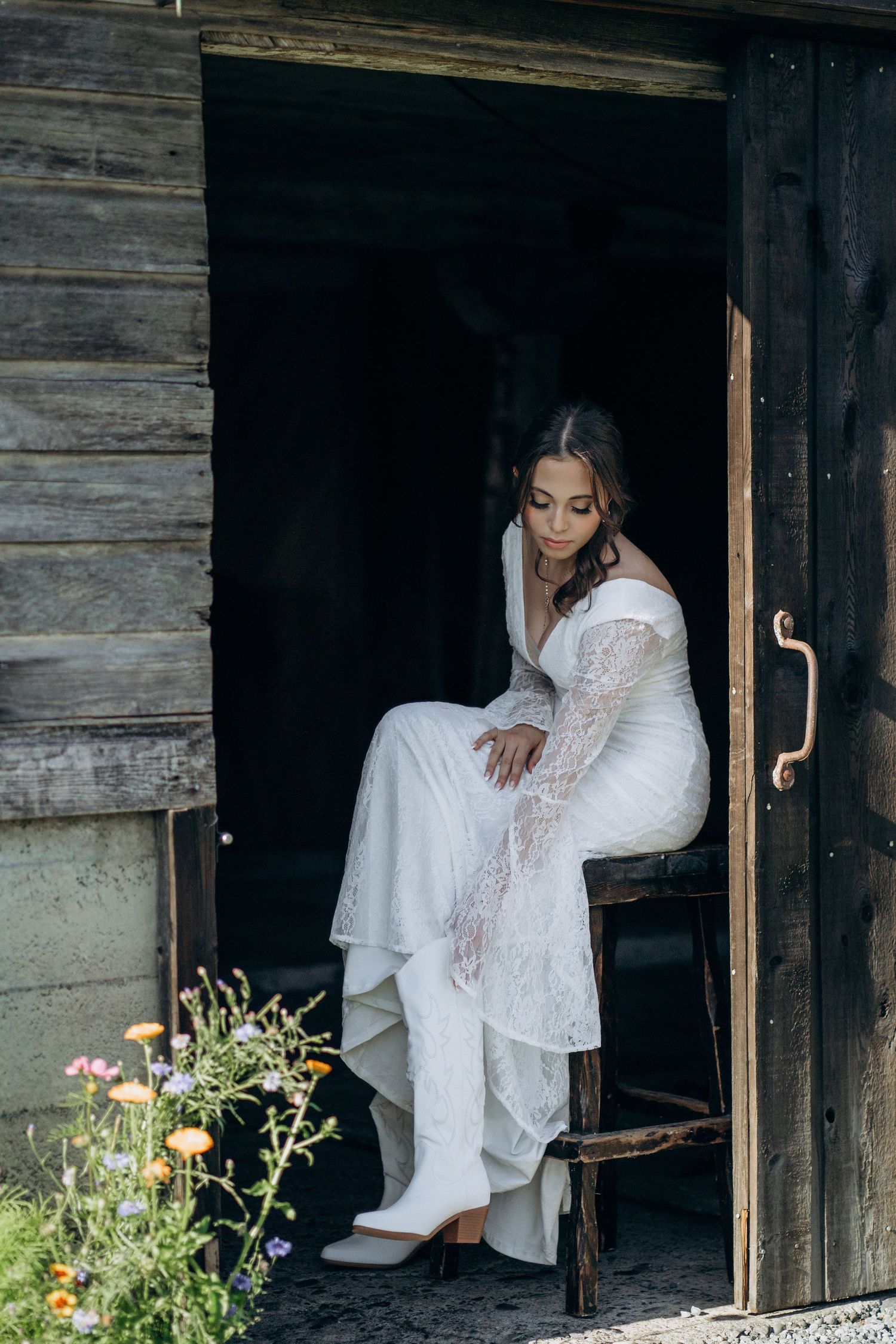 Bride in white lace wedding dress and boots seated in rustic barn doorway surrounded by wildflowers.