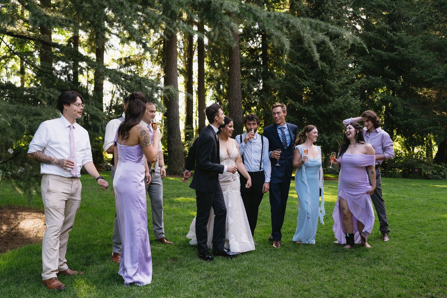 Wedding party in formal attire laughing and socializing on a lush green lawn surrounded by tall trees.