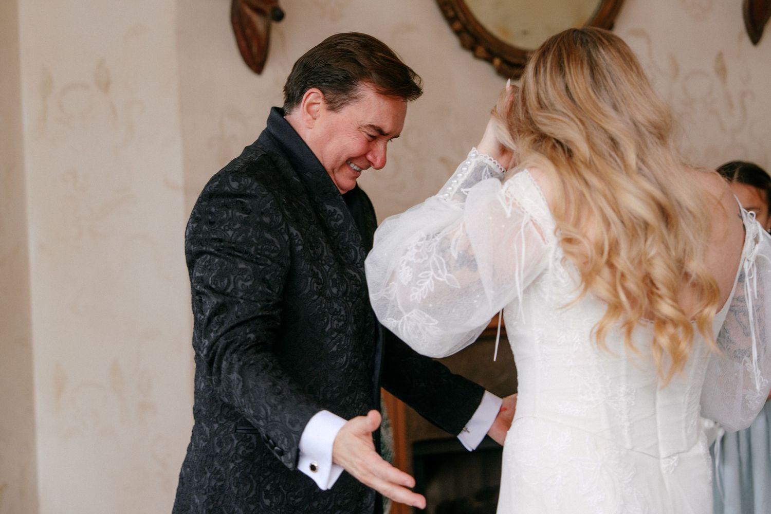 Groom in embroidered black jacket sharing an emotional first look reaction with his bride indoors.