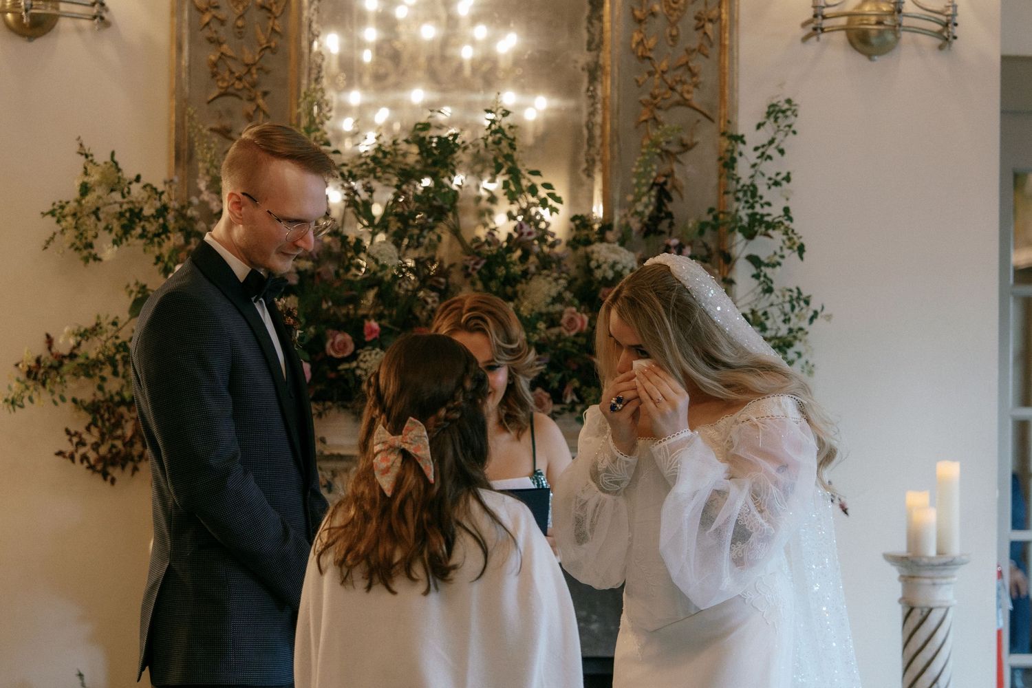 Bride wipes tears as groom and flower girl share tender moment at romantic indoor wedding.