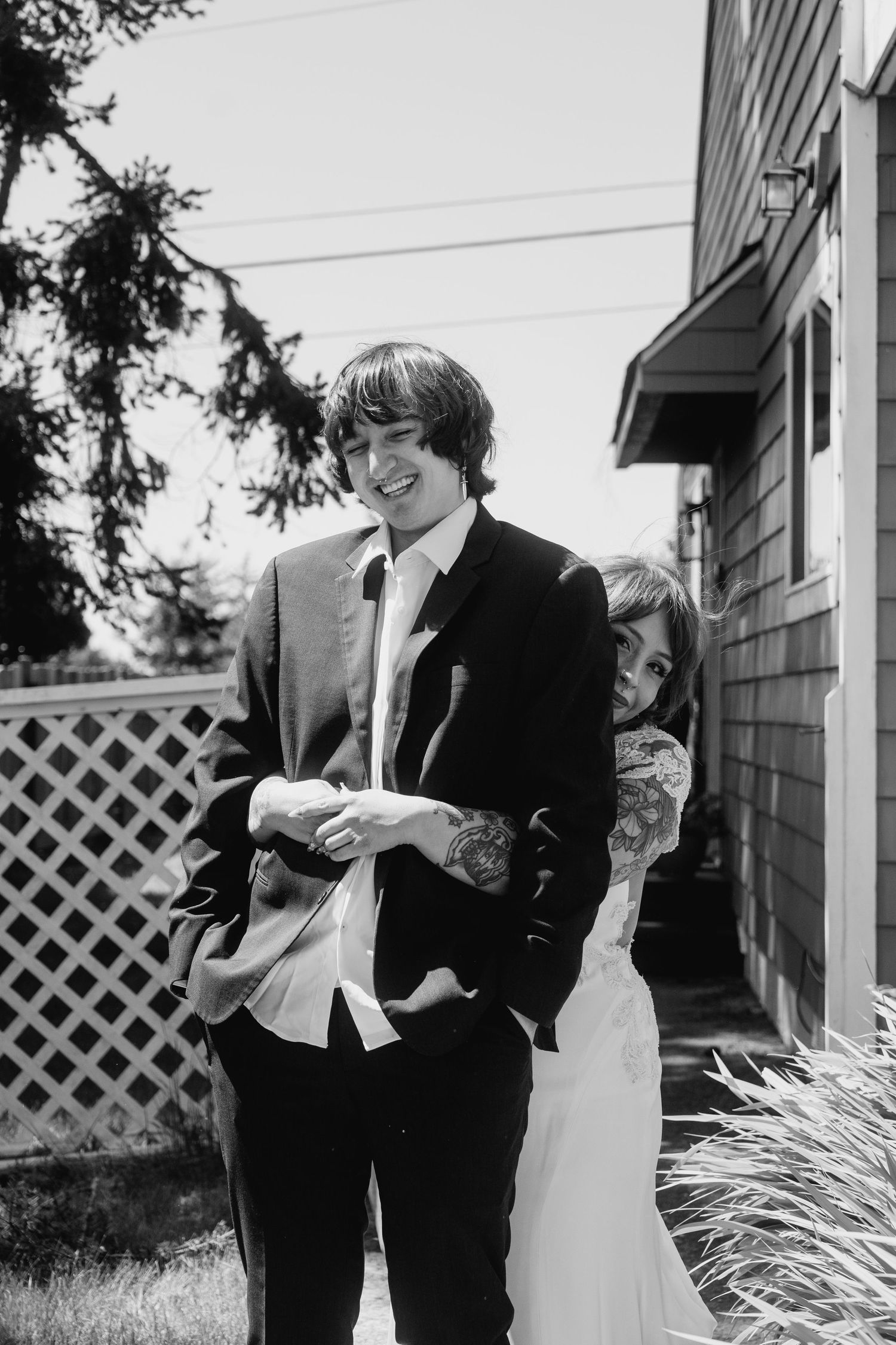Smiling groom in black suit stands outside as tattooed bride peeks playfully from behind him in black and white.