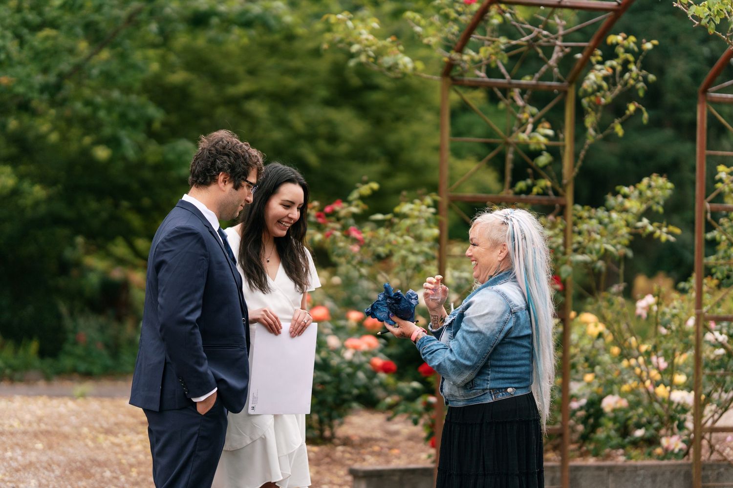 Bride and groom receive gift from older woman in jean jacket at outdoor rose garden wedding.