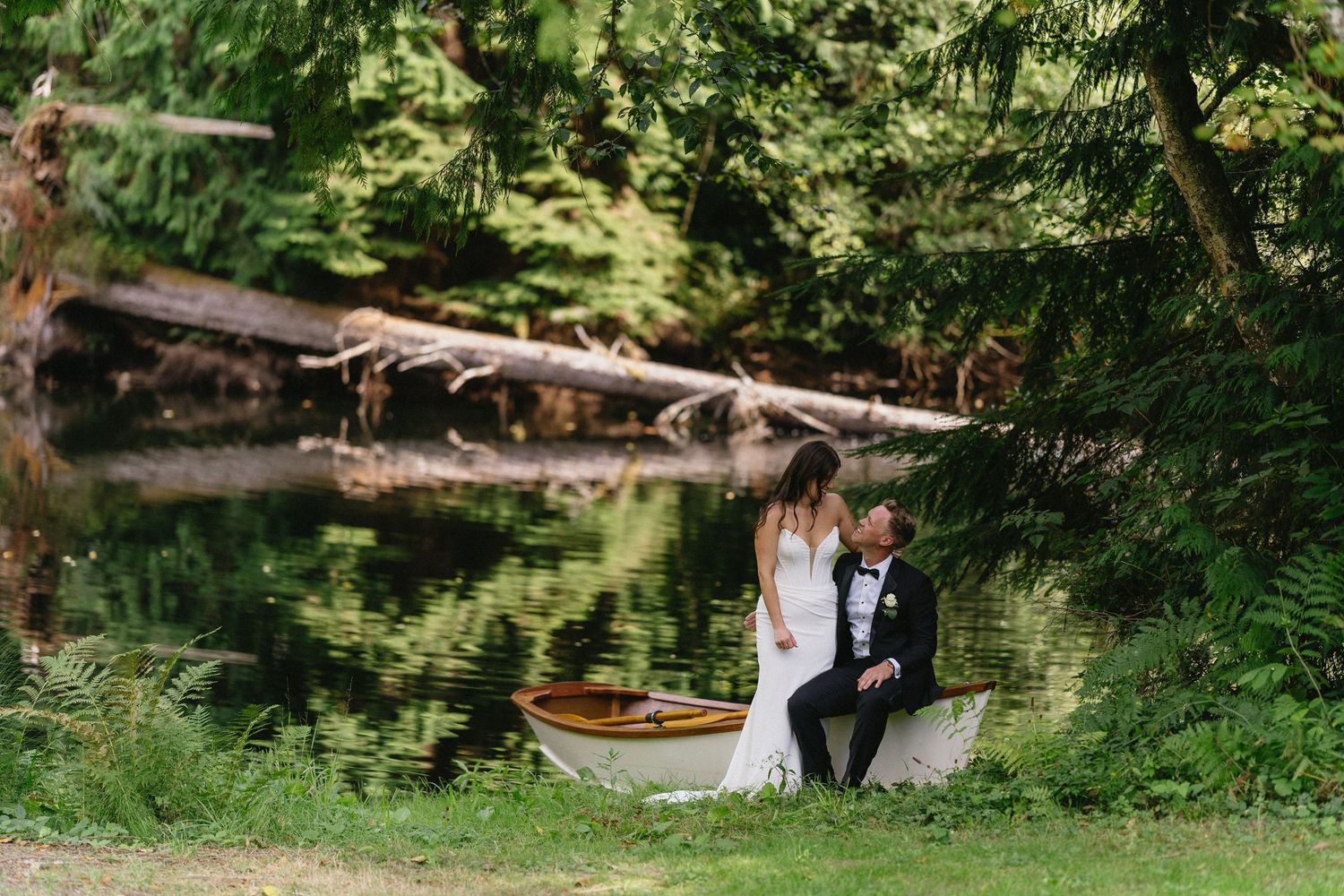 Wedding couple sitting by rowboat near serene forest pond reflection.