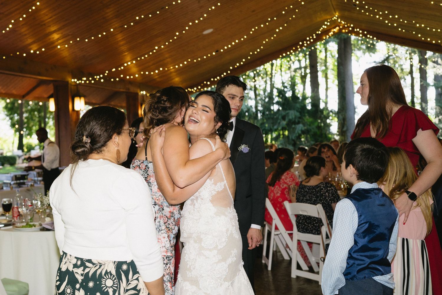 Bride in lace gown shares a joyful hug with a guest under twinkling fairy lights at reception.