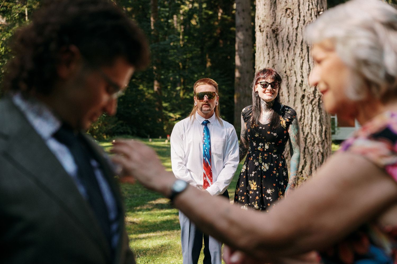 Man in white shirt with American flag tie smiling at outdoor wedding ceremony near large tree trunk.