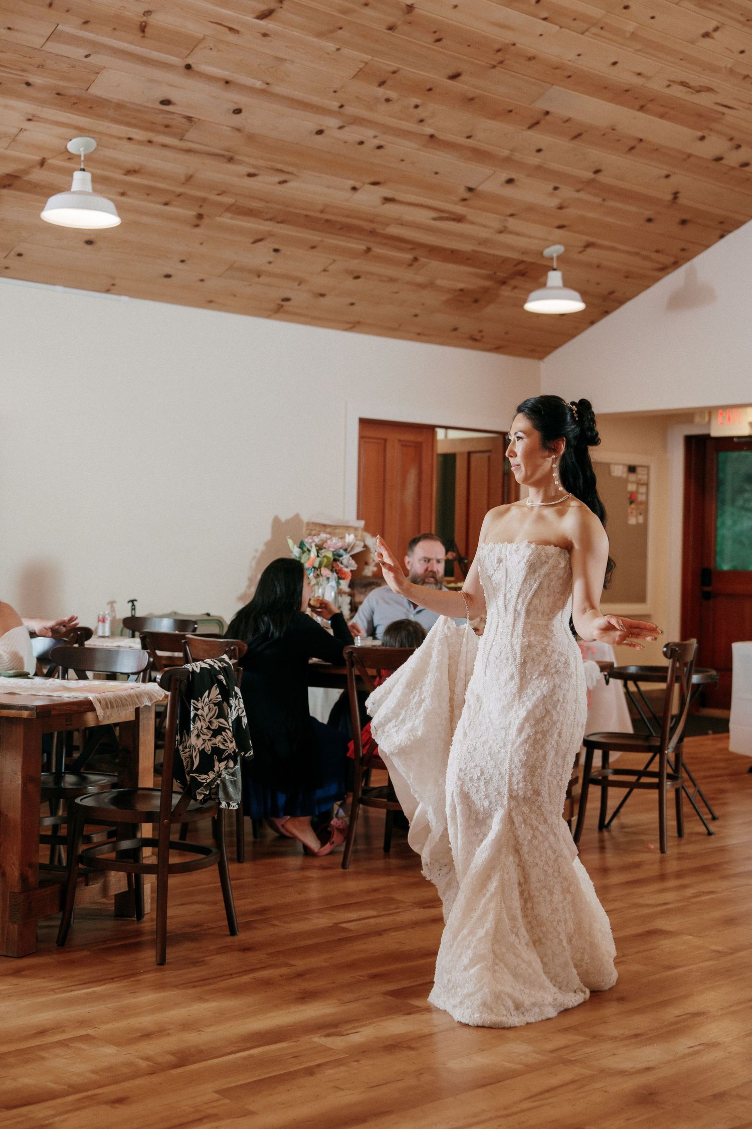 Bride in strapless lace gown twirling on hardwood floor inside rustic wooden-ceiling reception hall.