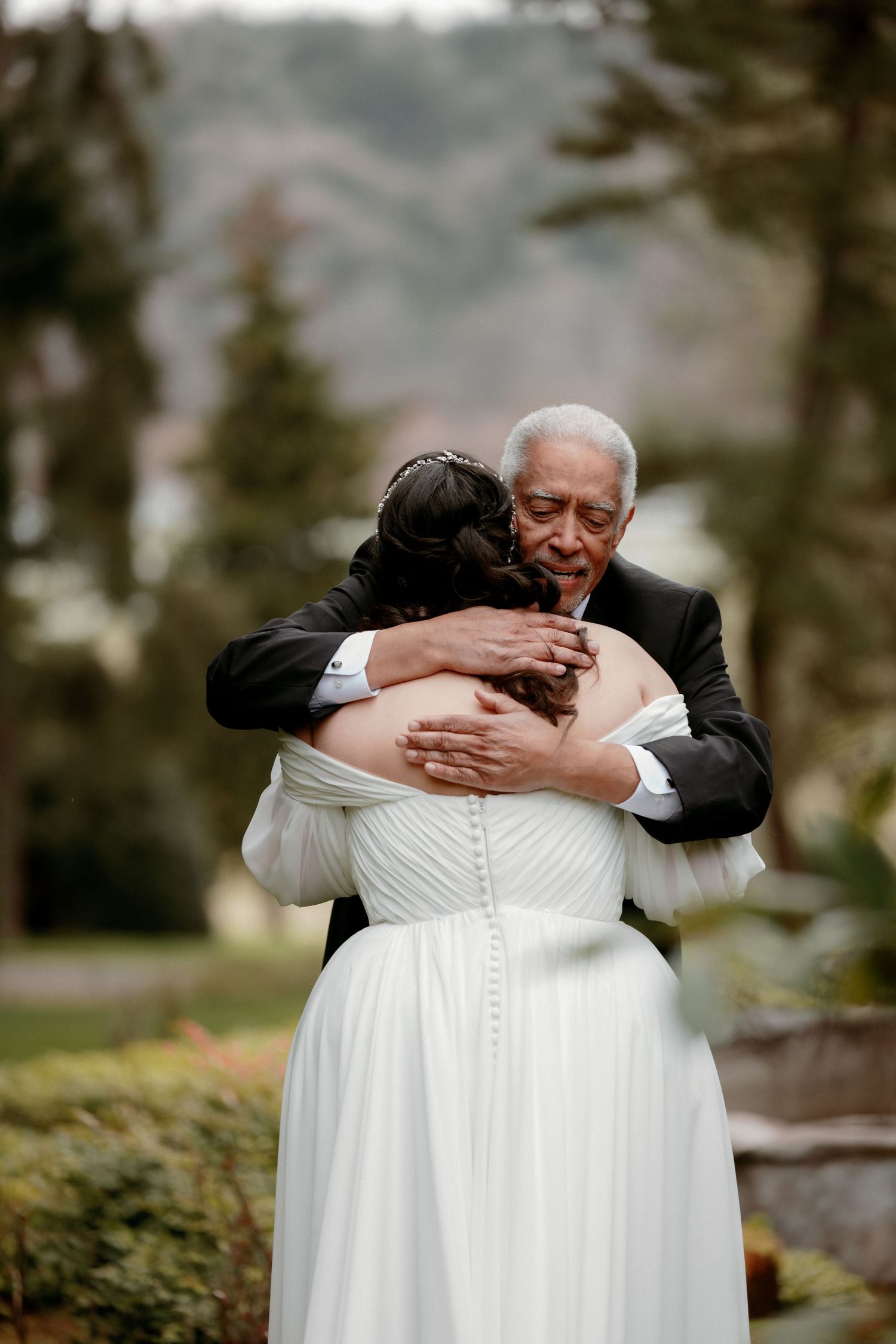 Elderly man in tuxedo sharing emotional hug with bride in white off-shoulder gown at outdoor wedding.