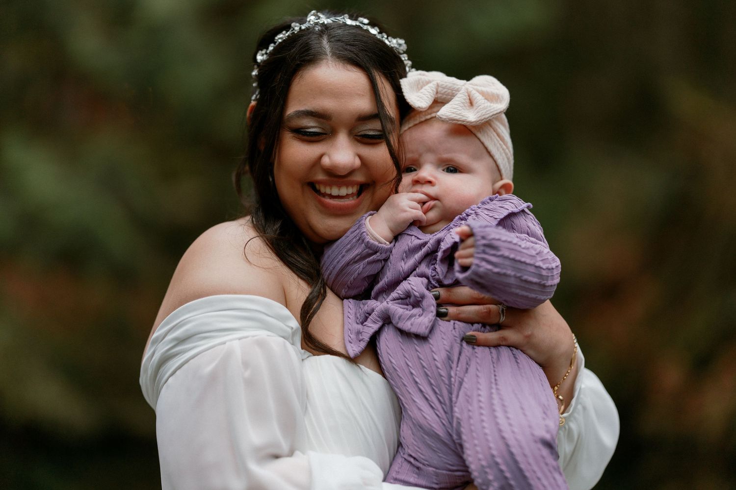 Smiling bride in white off-shoulder gown holding baby girl in purple outfit.