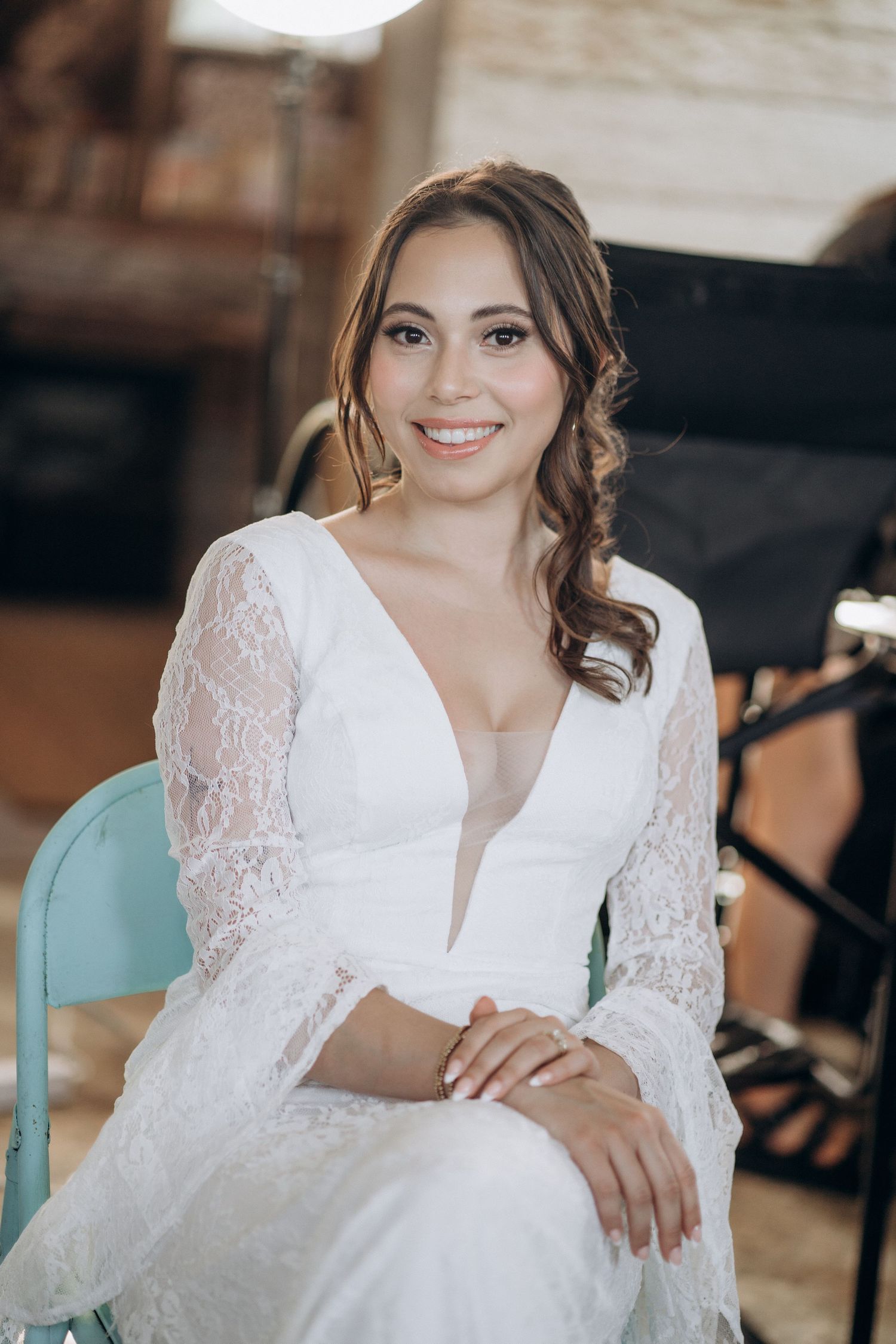 Smiling bride in white lace long-sleeve wedding dress seated on teal chair in rustic indoor setting.
