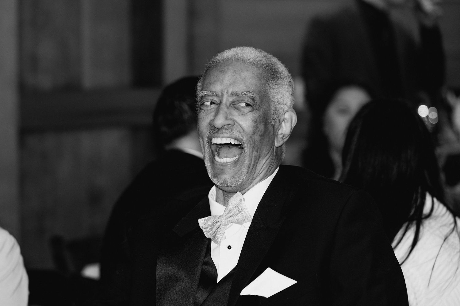 Elderly man in tuxedo laughing joyfully at a formal wedding reception in black and white photo.