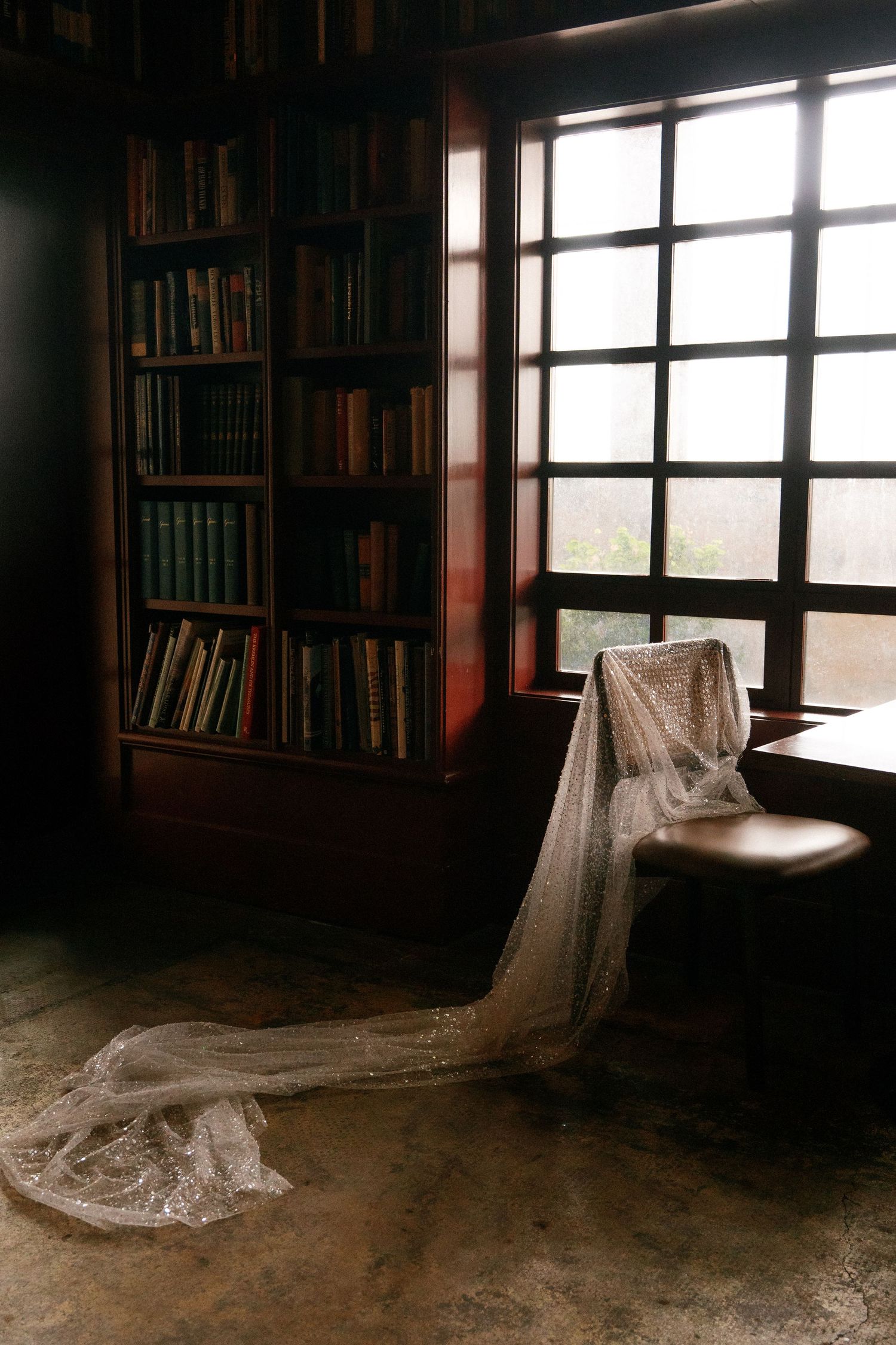 Elegant bridal veil draped over a leather chair beside a bookshelf and large window in a library.