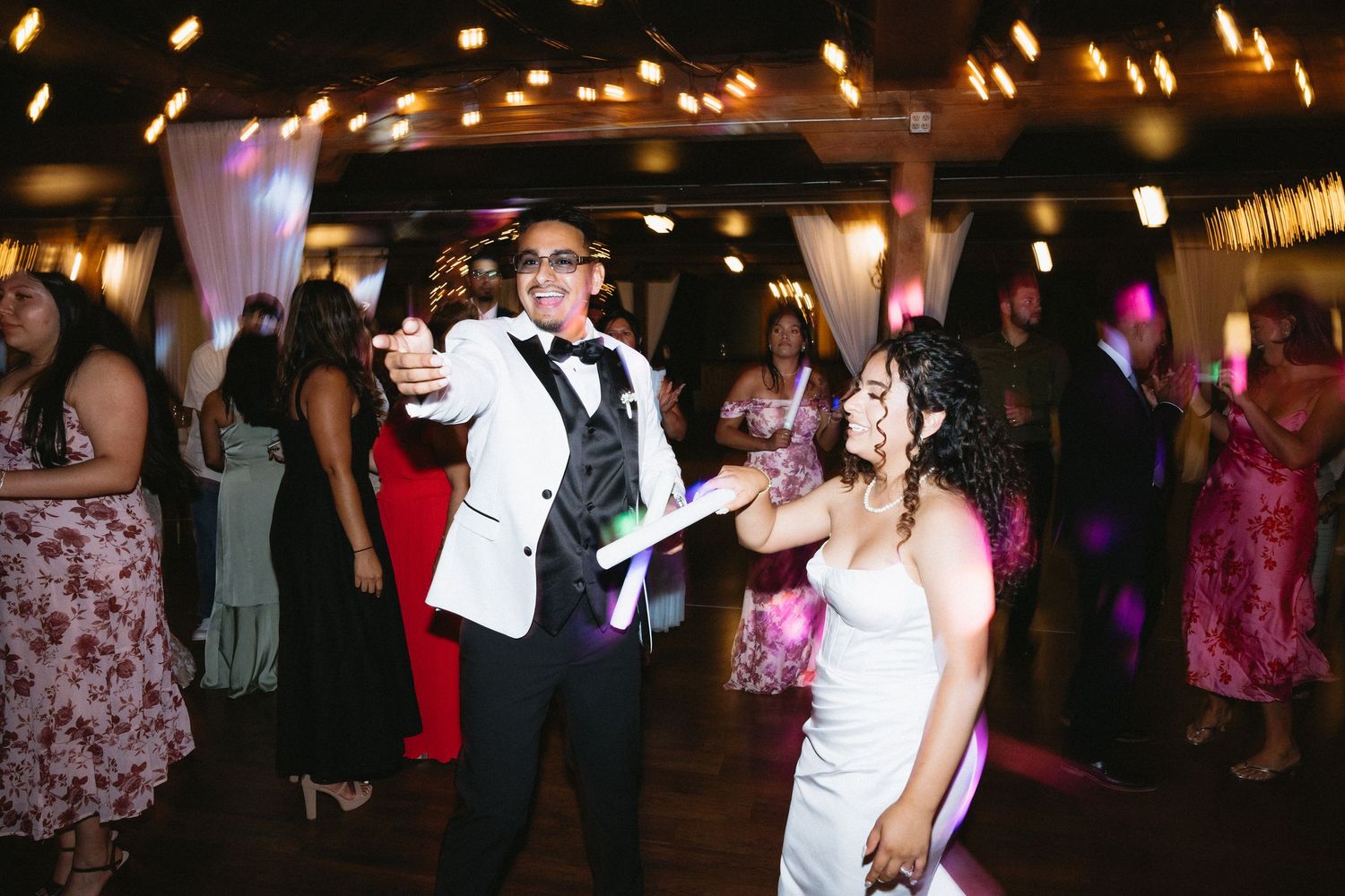 Bride and groom dance energetically with guests holding glow sticks at a vibrant wedding reception.