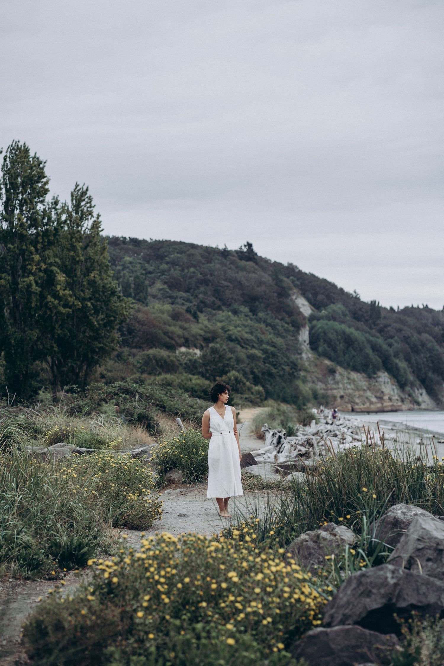 Woman in white dress standing on rocky coastal path with green hills and ocean in background.