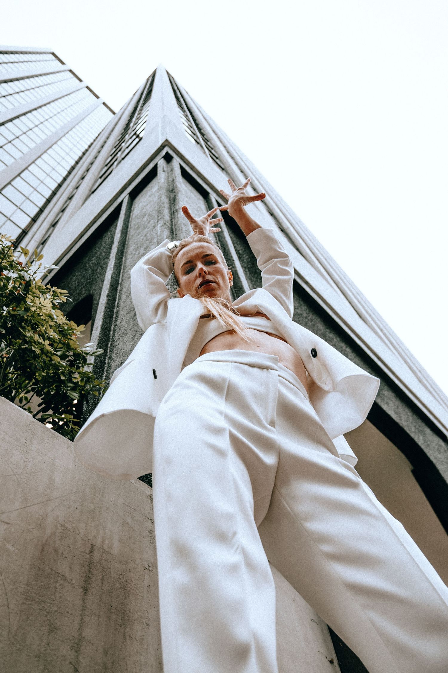 Low angle view of woman in white suit posing dramatically before tall glass skyscraper in city.