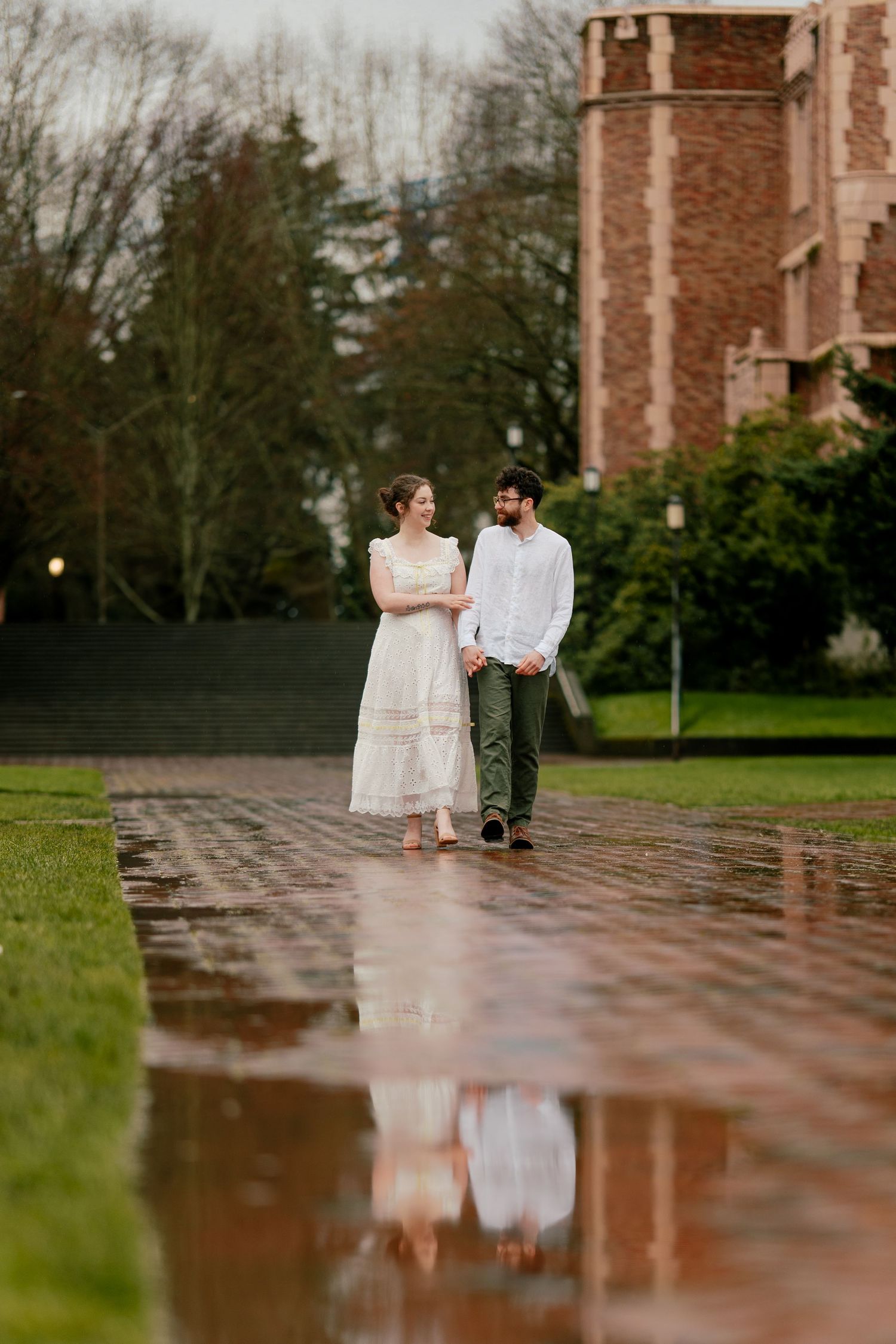 Couple walking hand in hand on a wet brick path with their reflection visible in a puddle.